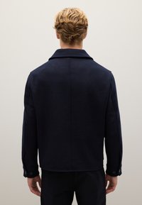 Giacca di lana blu navy con colletto, polsini abbottonati e vestibilità strutturata. Tessuto liscio e cuciture semplici visibili sul retro.
