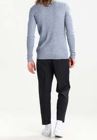 Personne portant un pull gris côtelé, un pantalon noir, des chaussettes blanches et des baskets blanches, se tenant de dos face à l'appareil photo sur un fond blanc.