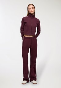 Donna in piedi che indossa una giacca a zip bordò e pantaloni della tuta abbinati con strisce bianche e sneaker bianche su uno sfondo neutro.