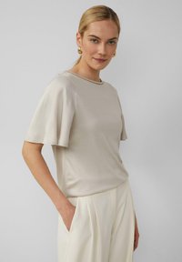 Beige blouse met korte mouwen, een gladde textuur, ronde hals en decoratieve stiksels bij de halslijn; gecombineerd met crèmekleurige broeken.