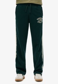 Donkergroene sweatpants met een trekkoord in de taille, zijstrepen in crème en de opdruk "Superdry JPN Athletic" aan de voorkant. Zacht, comfortabel materiaal.
