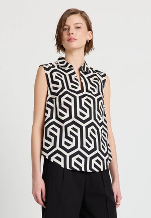 Donna che indossa una camicetta senza maniche con motivo geometrico bianco e nero e collo, abbinata a pantaloni neri, su uno sfondo chiaro e uniforme.