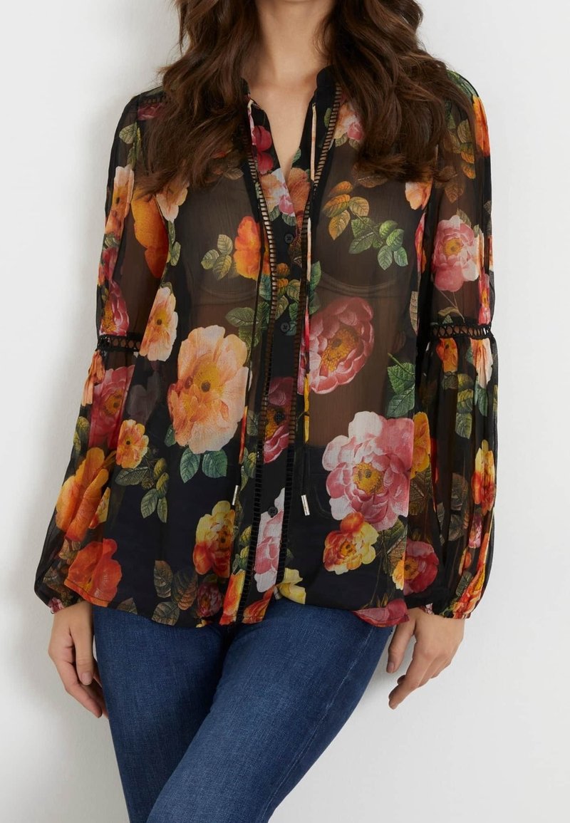 Femme portant un chemisier noir transparent avec des imprimés floraux orange vif, rose et jaune, manches longues et jean bleu.