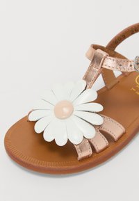 Sandal cu aur roz, decorat cu flori sintetice, petale albe și centru bej. Prezintă curea ajustabilă și talpă bej cu margine texturată.