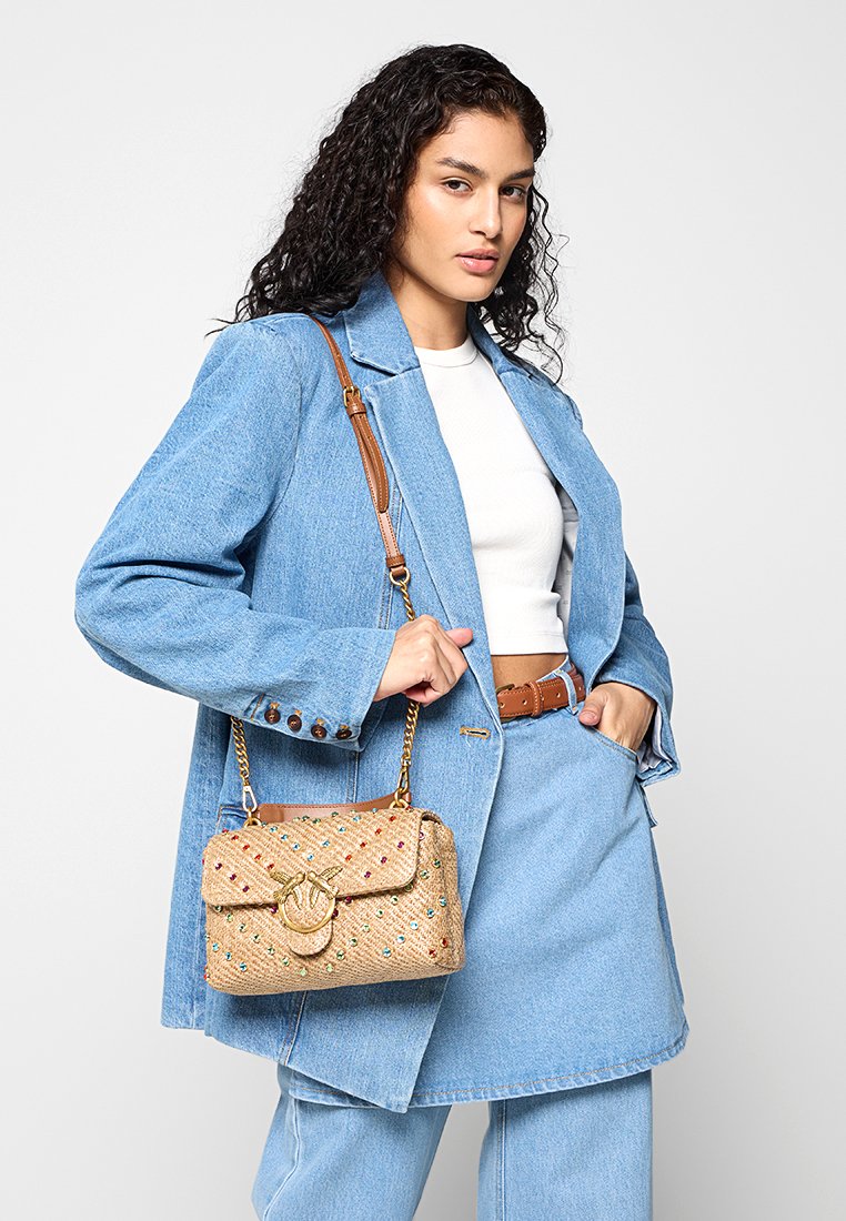 Mujer con cabello rizado y oscuro, vestida con una chaqueta de mezclilla clara y una falda, top corto blanco, cinturón marrón y llevando un bolso de hombro beige con tachuelas.