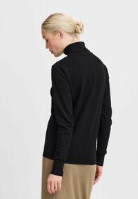 Pull en col roulé noir avec un design ajusté, col et poignets côtelés, texture lisse et silhouette simple, associé à un pantalon beige côtelé.
