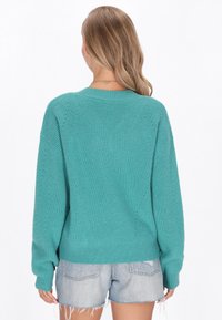 Teal kötött pulóver bordás textúrával, oversized szabással, lefittyedt vállakkal és hosszú ujjakkal, világos farmer rövidnadrággal párosítva.