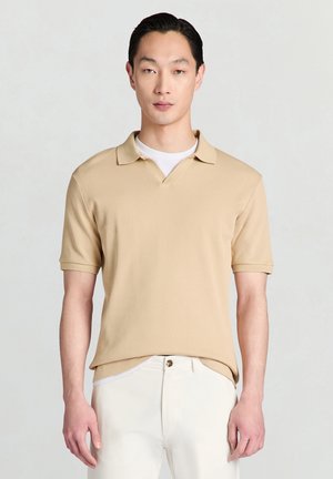 Giovane uomo con capelli pettinati all'indietro che indossa una polo beige a maniche corte a coste e pantaloni bianchi su uno sfondo chiaro e uniforme.