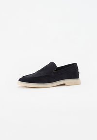  svarte semskede slip-on loafers med lys beige gummisåle på hvit bakgrunn, sidevinkel som viser sømdetaljer.