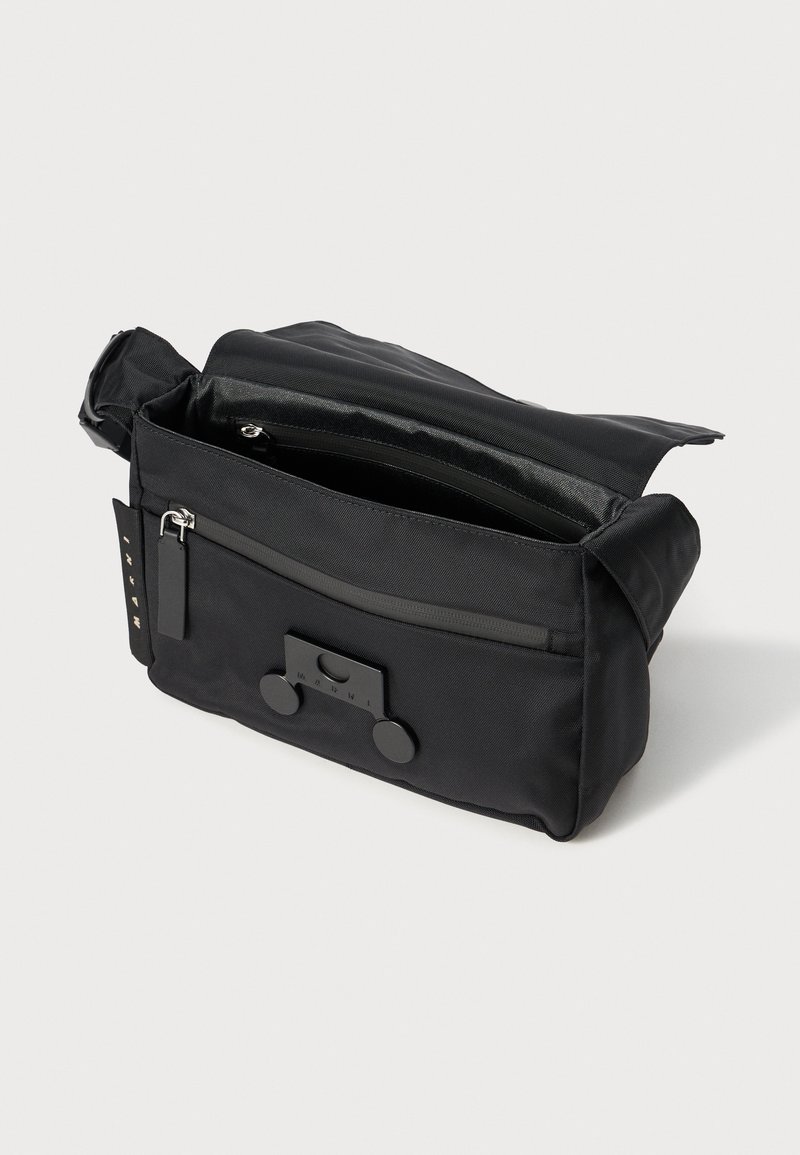 Schwarze Stofftasche mit Klappenverschluss, einer Front-Reißverschlusstasche, seitlichen Hardware-Akzenten und einem verstellbaren Tragegurt. Strukturierte Materialoberfläche.