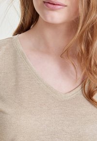 Ung kvinde med lys hud og langt rødt hår iført en beige V-hals skjorte, hvor hun viser nakke og delvist ansigt med lukkede læber.