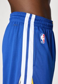 Blå basketballshorts laget av pustende mesh-materiale med hvite striper, elastisk midje, og et NBA-logo merke på siden.