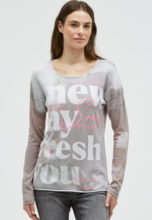 Young woman with long brown hair wearing a gray and white long-sleeve shirt with large pink and white text, paired with gray jeans.