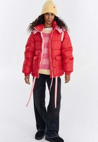 Czerwona kurtka puchowa z różowymi akcentami, zapinana na suwak z przodu, wyposażona w duże kieszenie i regulowane sznurki ściągające. Noszona na paskowanym swetrze z dzianiny i dżinsach.