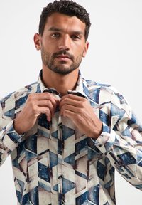 Man met kort, krullend haar en een baard die een geometrisch patroon gestructureerd shirt met blauwe, witte en bruine tinten dichtknopt tegen een effen achtergrond.