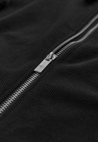 Black zip-up jacket featuring a textured fabric, silver metal zipper, and visible stitching along the edges of the zip area.