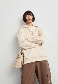 Pessoa com cabelo preto curto, vestindo um capuz bege da Adidas, calças cargo castanhas da Adidas e segurando uma bolsa bege.