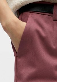 Burgunderfargede skreddersydde bukser med glatt tekstur og sidelomme, brukt med en kremfarget genser og svart belte synlig ved midjen.