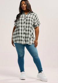 Camicia a maniche corte a quadri bianco e nero con chiusura a bottoni, abbinata a jeans skinny blu e sneaker bianche, che mostra un fit rilassato.