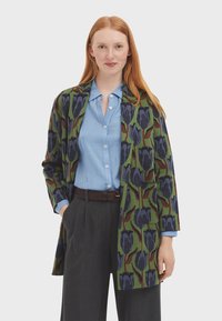 Cardigan vert à motifs floraux bleus et foncés, porté sur une chemise à col bleu clair, assorti à un pantalon à rayures.
