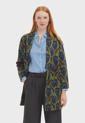Cardigan verde a fantasia con motivi floreali blu e scuri, indossato sopra una camicia colletto azzurra, abbinato a pantaloni a righe.