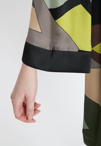 Vêtement à manches longues présentant un motif géométrique dans des tons de vert, de marron et de beige, avec un poignet noir au bas. Texture satinée et finition lisse.