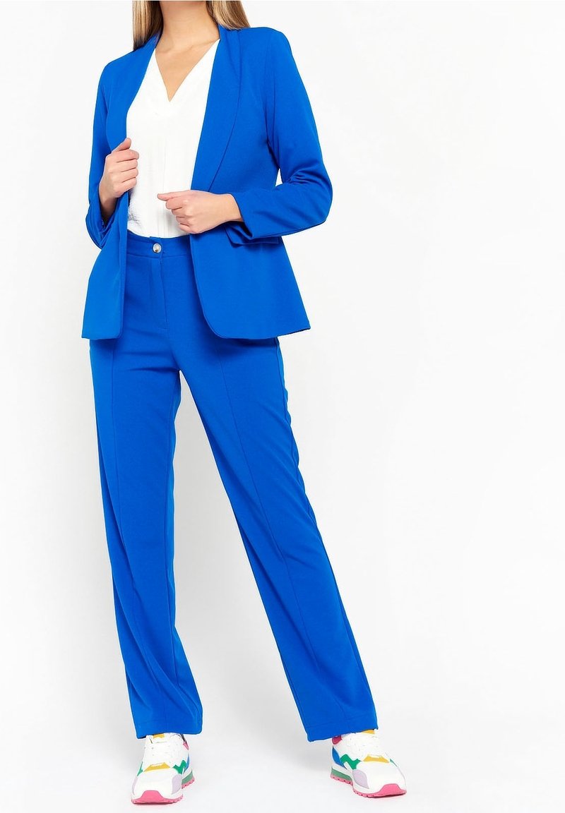 Femme portant un blazer et un pantalon bleu vif sur mesure, avec un top blanc à col en V et des baskets colorées, debout devant un fond blanc.
