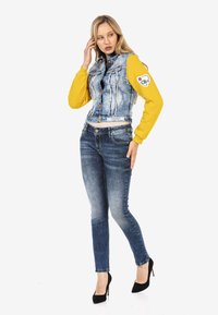 Gilet en denim avec des manches jaunes, fermetures à boutons à l'avant et détail de patch. Jean bleu ajusté avec effet usé et escarpins noirs.