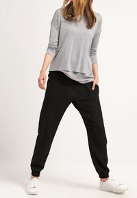 Nainen, joka pukeutuu harmaaseen pitkähihaiseen paitaan, mustiin breittyihinsuojiin ja valkoisiin lenkkareihin, seisoo jalat eristyksissä yksinkertaista taustaa vasten.