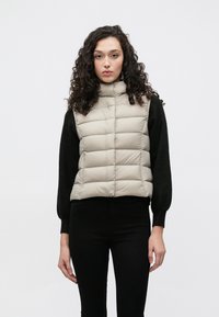 Vestă matlasată bej cu guler înalt, model matlasat, purtată peste un top cu mânecă lungă neagră, asortată cu pantaloni negri, pe un fundal neutru.