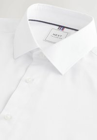 Bijela košulja s teksturiranim materijalom, klasičnim ovratnikom i malim dugmetom. Etiketa prikazuje "NEXT TAILORING" s obojenim naglascima.