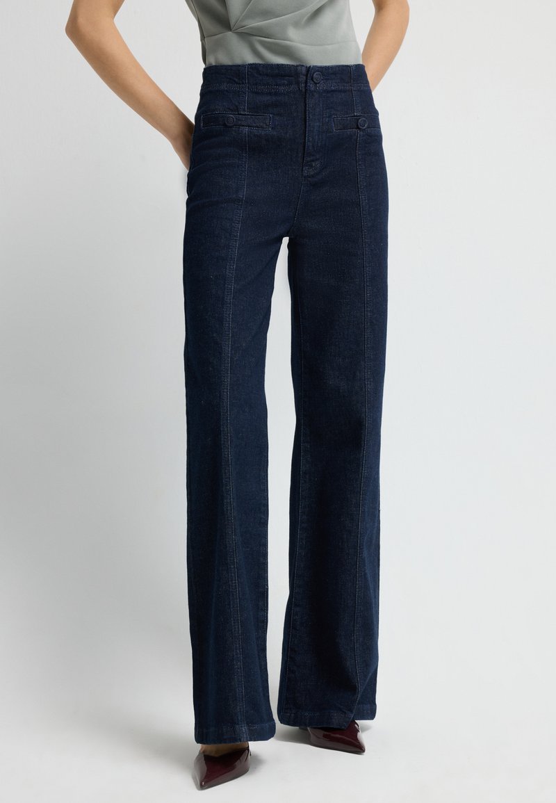 Tmavě modré riflové kalhoty s vysokým pasem a širokými nohavicemi, s předními kapsami a hladkým povrchem, doplněné špičatými vínovými podpatky.