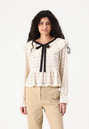 Blond bluse i off-white med rynket krage, svart båndtopp og peplum-bunn. Detaljert blomstermønster gjennom hele stoffet. Lange ermer.