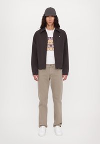 Jachetă neagră cu o croială lejeră, tricou grafic alb, pantaloni bej și adidași albi. Poartă o șapcă gri. Design simplu, fără modele proeminente.