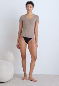Kortermet, tettsittende t-skjorte i tan, kombinert med svart høyutsnittet undertøy, vist på en modell i en nøytral setting. Mykt stoff, minimale sømmer.