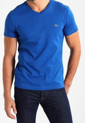 Mężczyzna ubrany w dopasowaną, niebieską koszulkę z dekoltem w serek i małym zielonym logo krokodyla na piersi oraz ciemnoniebieskie dżinsy, stojący z jedną ręką w kieszeni.