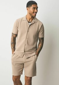 Beige two-piece short-sleeved shirt and shorts set, featuring a textured fabric with vertical stripes and a collar, buttoned front, and side pockets.