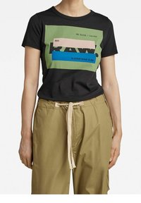 Person med en svart T-shirt med ett grafiskt block i grönt, rosa och blått samt beigea byxor med dragsko.