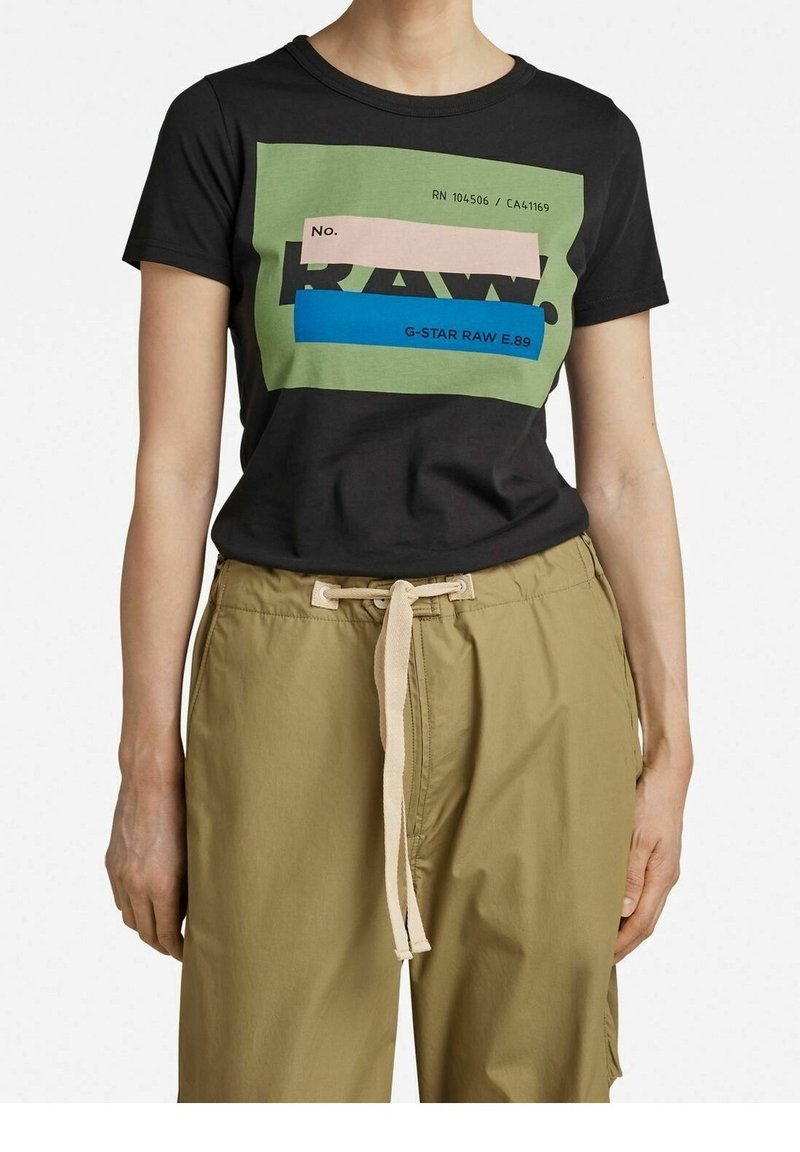 Person med en svart T-shirt med ett grafiskt block i grönt, rosa och blått samt beigea byxor med dragsko.