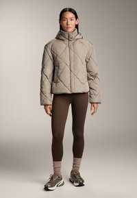 Giacca trapuntata beige con colletto alto, abbinata a leggings marroni e calze testurizzate, con scarpe da ginnastica dai dettagli grigi e neri.