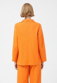 Chaqueta naranja con un cuello festoneado, hecha de una tela de textura suave. Presenta mangas largas y un corte entallado.