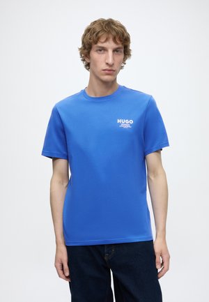 Jeune homme aux cheveux bouclés châtain clair portant un t-shirt bleu de la marque Hugo et un pantalon foncé, debout devant un fond clair uni.