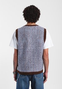 Gilet en tricot avec des rayures verticales bleues et brunes, doté d'une bordure brune au niveau de l'encolure et de l'ourlet. Porté par-dessus un t-shirt blanc et un jean en denim.