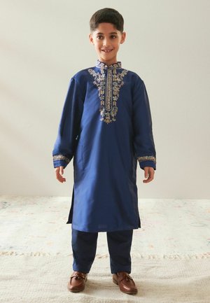 Dreng står smilende iført en blå traditionel dragt med guldbroderi, brune sko, foran en ensfarvet lys baggrund.