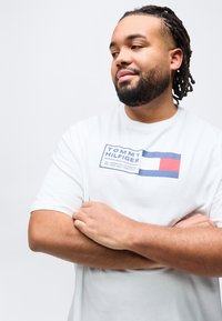 Άνδρας με γενειάδα και dreadlocks, φορώντας λευκό μπλουζάκι Tommy Hilfiger, με σταυρωμένα τα χέρια, κοιτάζει προς το πλάι σε απλό λευκό φόντο.