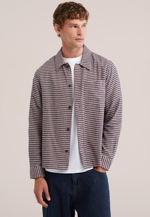 Mann trägt eine violett-beige Hahnentritt-Knopfleiste Jacke über einem weißen Hemd und dunkelblauen Jeans, schaut zur Seite.