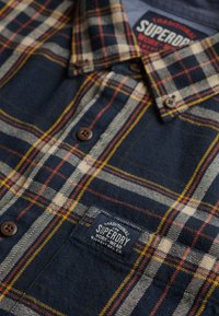 Camisa de franela con un patrón de cuadros en azul marino, beige y rojo, con botones marrones y una etiqueta azul que dice "Superdry Traditional Workwear".