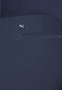 Navy blue athletic pants featuring a smooth, stretchy texture, a small black logo tag, and a discreet pocket detail on the back.