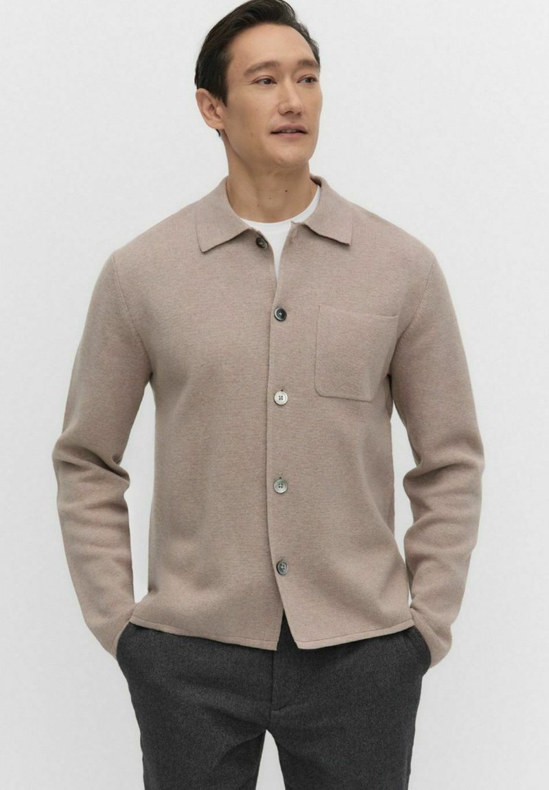 Lichtbeige cardigan van zachte stof, met een spitse kraag, knopen aan de voorkant en een enkele borstzak. Effen kleur, minimalistisch ontwerp.