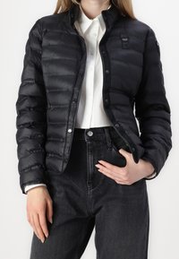 Sort polstret jakke med trykknap-lukning, med en klassisk krave og minimal branding. Båret over en hvid skjorte og mørke jeans.